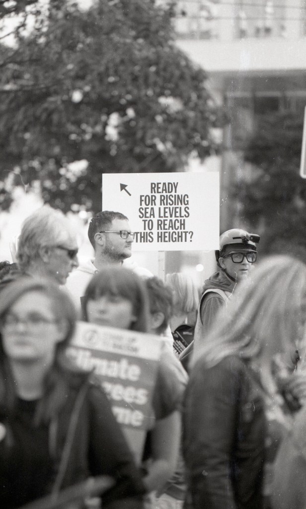 image description: taken through a crowd, in between two people we see a placard depicting the height that rising sea levels will reach due to climate change. text reads: "ready for rising sea levels to reach this height?"
